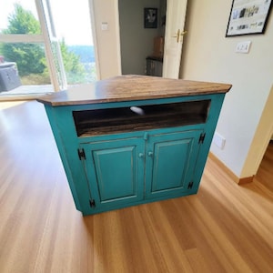 May include: A teal-colored corner cabinet with a wooden top. The cabinet has two doors and a shelf. The doors have black hinges and handles. The cabinet is made of wood and has a distressed finish.