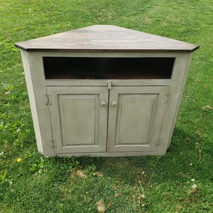May include: A corner cabinet with a triangular top and a light gray painted finish. The cabinet has two doors with white knobs and a dark brown top. The cabinet is designed to fit into a corner space.