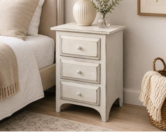 May include: A white wooden nightstand featuring three drawers with small round knobs and a distressed finish. The rectangular piece stands on four arched legs and includes a flat top surface. The design incorporates a frame-and-panel construction.