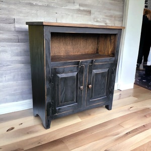 May include: A black wooden cabinet with two doors and a shelf. The cabinet has a dark finish with visible wood grain and a lighter brown top. The doors have wooden knobs and decorative hinges. The cabinet is on a light wood floor.