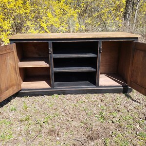 A black and brown wooden cabinet with three doors. The doors are open and reveal shelves inside. The cabinet is sitting on a patch of grass.