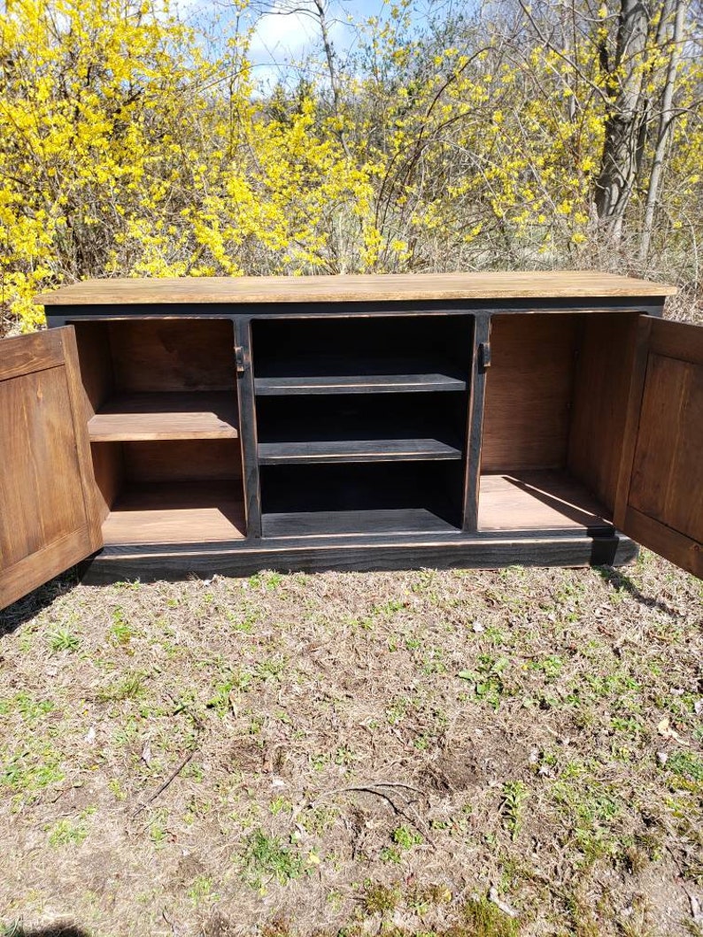 A black and brown wooden cabinet with three doors. The doors are open and reveal shelves inside. The cabinet is sitting on a patch of grass.