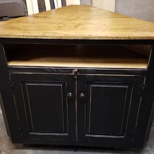 May include: A corner cabinet with a light brown, triangular top and black base. The cabinet has two doors and an open shelf. The cabinet is made of wood and has a distressed finish. The cabinet is designed to fit in a corner.