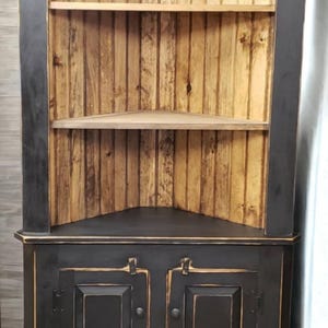 May include: A black corner cabinet with a distressed finish. The upper section has open shelves with a natural wood backing and shelves. The lower section features two doors with knobs. The cabinet is designed for corner placement.