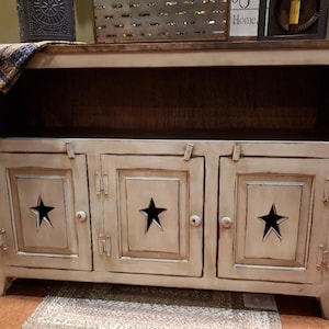 May include: A cream-colored wooden cabinet with three doors, each featuring a cut-out star design. The cabinet has a dark brown top and a distressed finish, with metal hardware and a shelf in the middle. The cabinet is a rustic style.