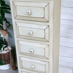 May include: A white wooden chest of drawers with five drawers and white knobs. The drawers have a distressed finish and the chest has a slightly curved base.