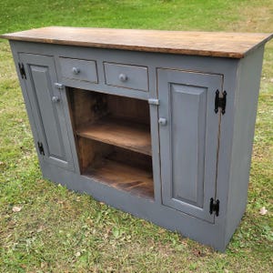 May include: A gray painted wooden cabinet with two doors, three drawers, and two shelves. The cabinet has black metal hinges and knobs. The top of the cabinet is made of a darker wood.
