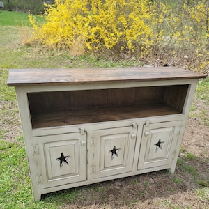 May include: A distressed, off-white wooden cabinet with a dark brown top. The cabinet has three doors, each featuring a cut-out star design. The cabinet is set outdoors on a grassy area, with yellow flowers in the background.