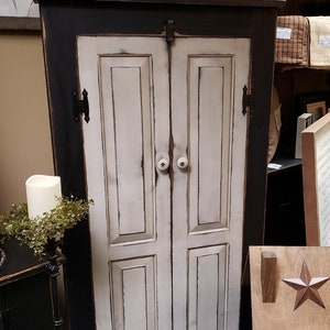 May include: A black and white distressed cabinet with two doors and white knobs. The cabinet has a distressed finish. On top are three white jars with the words "Home Sweet Home" and greenery.