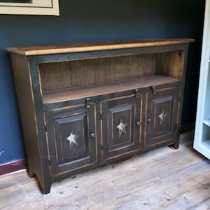 May include: A dark-stained wooden cabinet with three doors and an open shelf. Each door features a cut-out star design. The cabinet has a natural wood top and is set against a dark blue wall.
