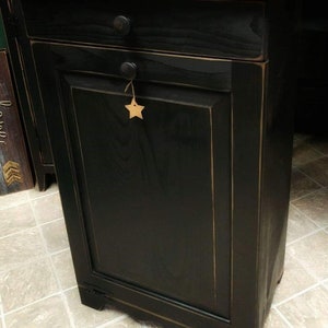 May include: Black wooden trash can with a single drawer and a star-shaped tag. The can has a distressed finish and a wood top.