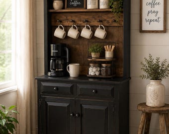 May include: A black wooden hutch with two drawers and two cabinet doors, featuring an open shelf with four hooks holding white mugs, a coffee maker, and a two-tiered stand with jars. The hutch includes signs reading but first, coffee, COFFEE, SUGAR, and coffee pray repeat.