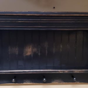 May include: Black wooden wall shelf with a decorative top and a lower shelf with hooks. The shelf has a distressed finish.