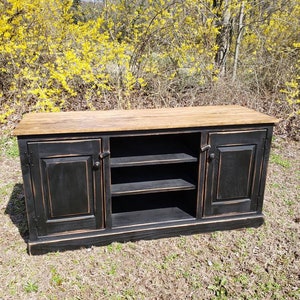 A black wooden media console with two doors and two shelves. The console has a distressed finish and a natural wood top. The console is sitting on a grassy surface.