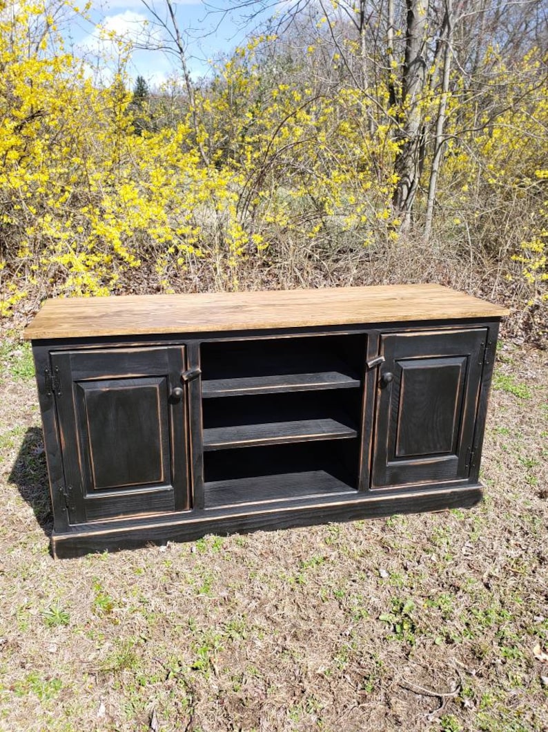 A black wooden media console with two doors and two shelves. The console has a distressed finish and a natural wood top. The console is sitting on a grassy surface.