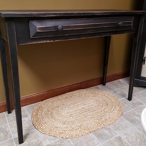 May include: A black wooden console table with a single drawer and round knobs. The table has a distressed finish and sits on four slender legs. An oval jute rug is on the floor in front of the table.