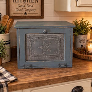 May include: A rectangular blue wooden bread box featuring a hinged front door with a central metal panel. The metal panel displays a punched star design surrounded by a decorative border. A small round knob is centered at the top of the door.