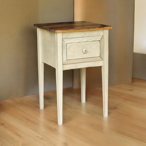 May include: A small, off-white wooden nightstand with a single drawer and a dark brown top. The nightstand has a square top, a drawer with a round knob, and four slender legs. The nightstand is approximately 24 inches tall.