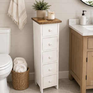 May include: A tall, narrow white storage cabinet featuring five stacked drawers with small round white knobs and a dark brown stained wooden top surface. The unit stands on four short legs and is positioned in a bathroom setting.