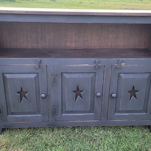 May include: A dark gray wooden cabinet with three doors, each featuring a star cutout. The cabinet has a shelf and a lighter-colored top. The doors have metal latches and hinges. The cabinet is on a grassy surface.