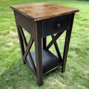 May include: A wooden end table featuring a natural brown stained rectangular top and a black painted base. The piece includes a single drawer with a small round black knob and an open lower shelf. The sides are designed with a structural X-frame.