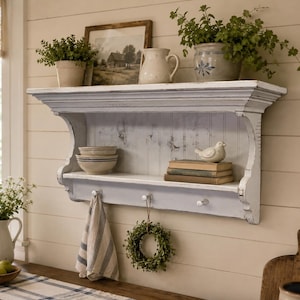 May include: A white wall-mounted wooden shelf with a distressed finish, featuring a top ledge for decor, a middle shelf holding bowls and books, and three pegs underneath. A striped towel and a small green wreath hang from the pegs.