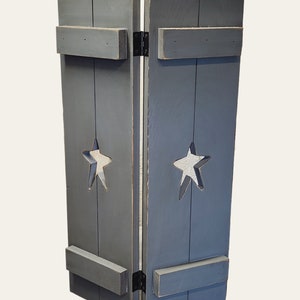 May include: A two-panel, gray wooden room divider with star cutouts. The panels are connected by black hinges and feature horizontal wooden supports. The star cutouts are centered on each panel, adding a decorative touch to this rustic home decor item.