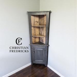 May include: A dark gray corner cabinet with a wooden interior and two shelves. The cabinet has a single door and is placed in a corner against a gray wall. The text "CHRISTIAN FREDRICKS" is visible.
