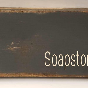 A rectangular wooden sign with a dark gray painted surface and the word 'Soapstone' written in white paint.