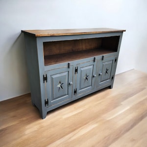 May include: A gray wooden cabinet with three doors and a star cut-out design. The cabinet has a brown wooden top and a shelf. The cabinet is painted gray with black hinges and hardware. The cabinet is sitting on a wooden floor.
