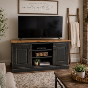 May include: A black wooden media console with a natural wood-toned top, featuring two side cabinets and three central open shelves. The console supports a flat-screen television and is positioned below a sign that reads AS FOR ME & MY HOUSE.