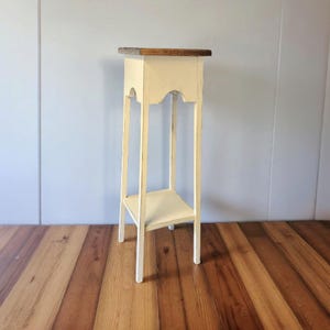 May include: A tall, cream-colored plant stand with a rectangular top and a lower shelf. The top is stained a dark brown. The stand has a distressed, vintage look, with decorative cutouts on the sides. The stand is approximately 30 inches tall.