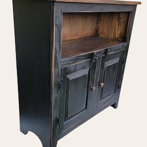 A black wooden cabinet with two doors and a shelf. The cabinet has a distressed finish and a simple design. The doors have a raised panel design and are accented with black metal hardware. The shelf is made of wood and is visible through the open doors.