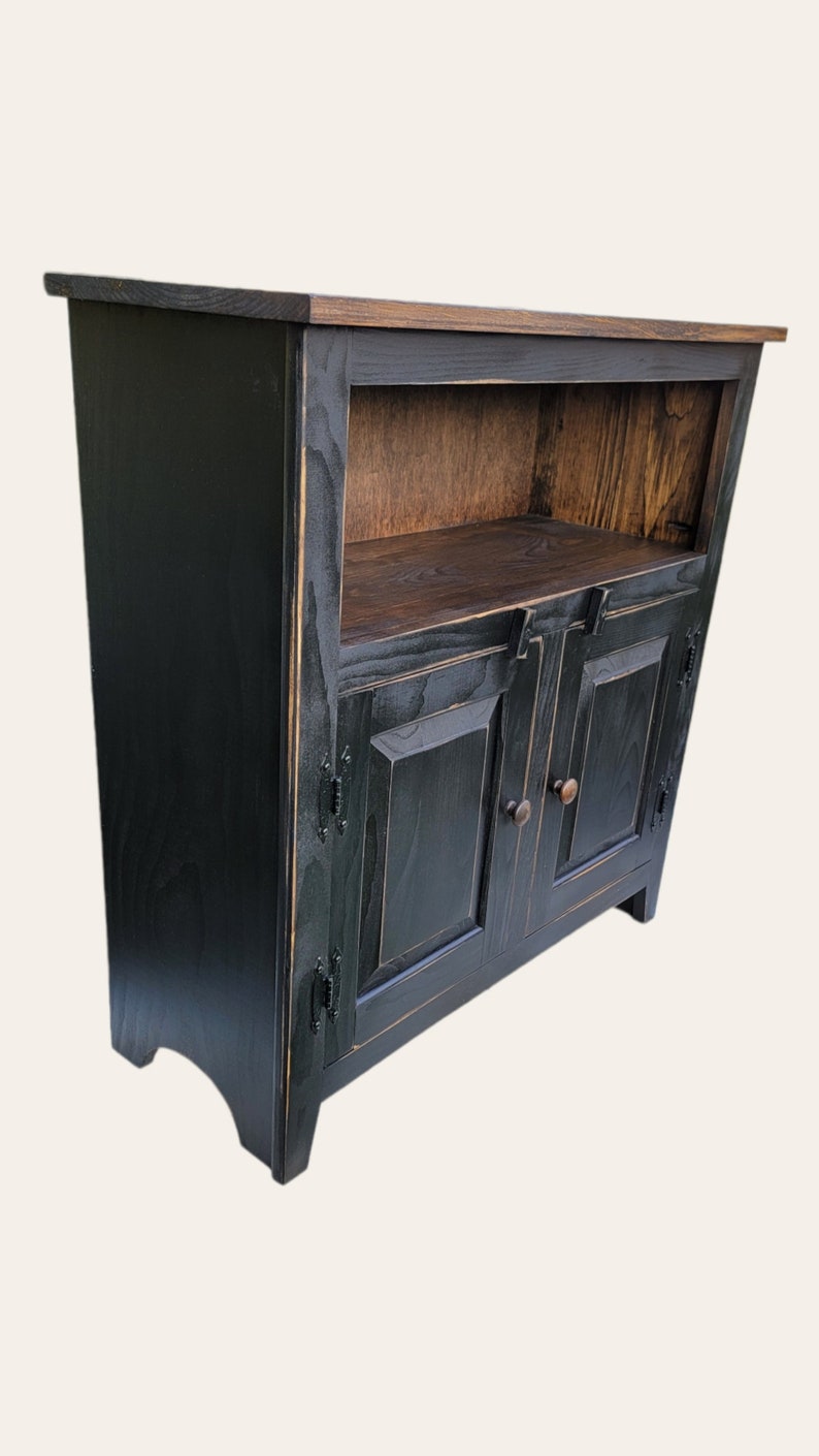 A black wooden cabinet with two doors and a shelf. The cabinet has a distressed finish and a simple design. The doors have a raised panel design and are accented with black metal hardware. The shelf is made of wood and is visible through the open doors.
