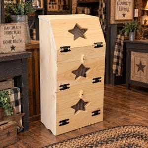 May include: A light-colored wooden storage bin featuring three stacked compartments, each with a star-shaped cutout covered by wire mesh. The unit has black metal hinges on the doors and small wooden knobs for opening the lower two sections.