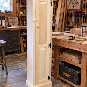 May include: A tall, narrow, light-colored wooden cabinet featuring a single door with two recessed panels, a small round wooden knob, and three black metal hinges. The cabinet has a flat top and a base, standing upright in a workshop setting.