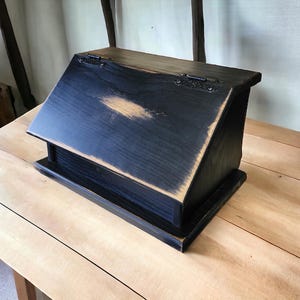 May include: A black wooden box with a slanted top and a hinged lid. The box has a distressed finish, revealing the wood grain in places. The box is sitting on a light-colored wooden surface.
