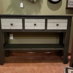May include: A dark gray console table with three drawers and a lower shelf. The drawer fronts are light beige with dark knobs. The table has a rectangular top and a distressed finish, suitable for a living room or entryway.