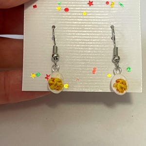 以下が含まれることがあります： シルバーカラーのフックが付いたぶら下がりピアス。各イヤリングには、黄色とオレンジ色のデザインが施された小さな白い楕円形のチャームが付いています。イヤリングは、カラフルな紙吹雪で飾られた白いカードに表示されています。