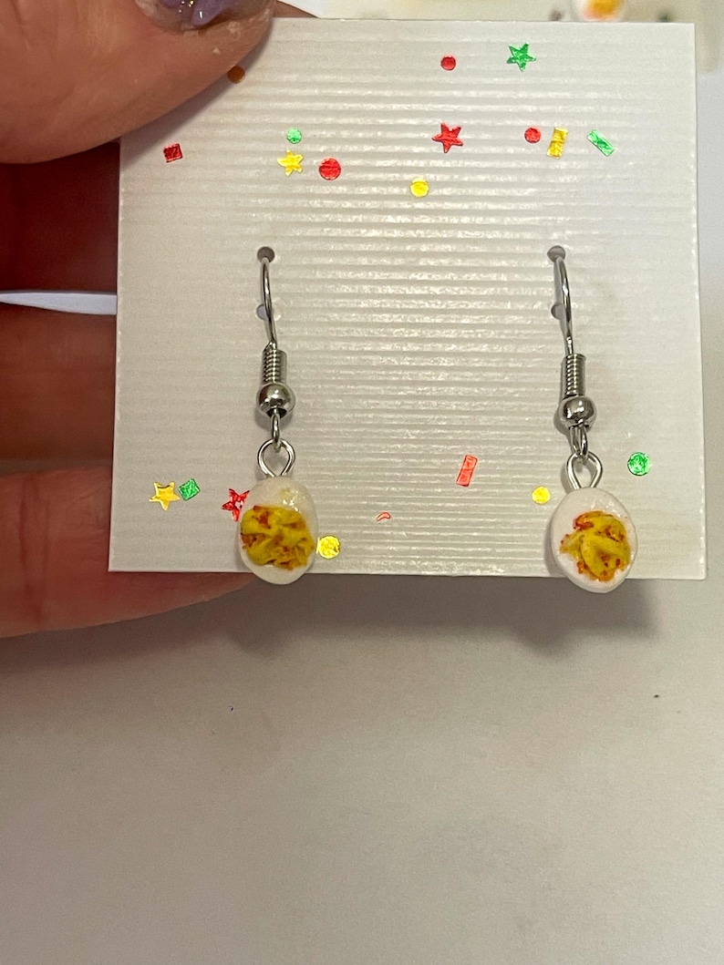 以下が含まれることがあります： シルバーカラーのフックが付いたぶら下がりピアス。各イヤリングには、黄色とオレンジ色のデザインが施された小さな白い楕円形のチャームが付いています。イヤリングは、カラフルな紙吹雪で飾られた白いカードに表示されています。