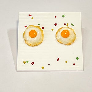 May include: A pair of earrings shaped like fried eggs with yellow yolks and white egg whites. The earrings are on a white surface with red, green, and yellow confetti.