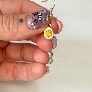 May include: A single silver earring with a white, egg-shaped charm. The charm is decorated with yellow and orange details, resembling a sliced hard-boiled egg. The earring is held by a silver hook.