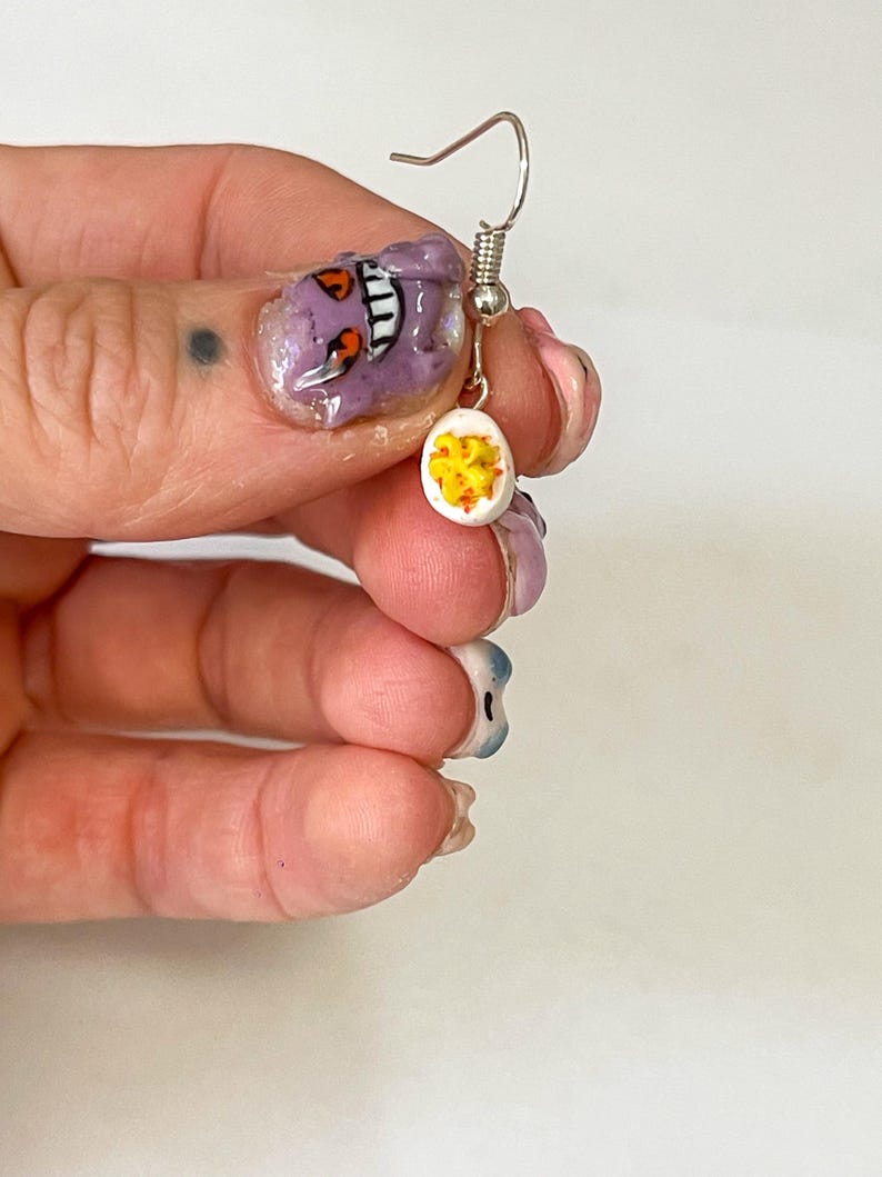 以下が含まれることがあります： 白い卵型のチャームが付いた、片方のシルバーのイヤリング。チャームは、スライスしたゆで卵に似た黄色とオレンジ色のディテールで装飾されています。イヤリングはシルバーのフックで吊り下げられています。