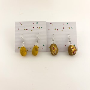 May include: A pair of dangle earrings featuring miniature food charms. One pair is a yellow potato and the other is a hot dog with mustard and ketchup.