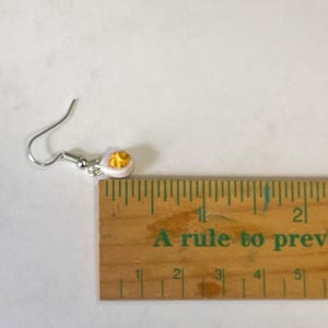 以下が含まれることがあります： 白と黄色の目玉焼きのデザインのシングルイヤリング。イヤリングはシルバーのフィッシュフックに取り付けられています。「A rule to prev」というテキストが入った木製の定規がイヤリングの隣にあります。
