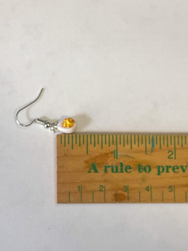以下が含まれることがあります： 白と黄色の目玉焼きのデザインのシングルイヤリング。イヤリングはシルバーのフィッシュフックに取り付けられています。「A rule to prev」というテキストが入った木製の定規がイヤリングの隣にあります。