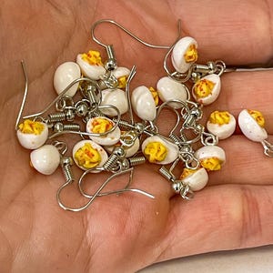以下が含まれることがあります： 銀色のフックピアスが付いた、ミニチュアのゆで卵風イヤリングのコレクション。イヤリングは、黄色の卵黄が詰められた白い卵の半分を特徴とし、リアルな食品をテーマにしたアクセサリーを作成しています。