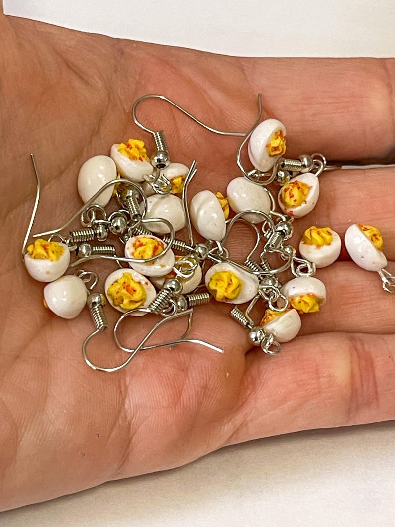 以下が含まれることがあります： 銀色のフックピアスが付いた、ミニチュアのゆで卵風イヤリングのコレクション。イヤリングは、黄色の卵黄が詰められた白い卵の半分を特徴とし、リアルな食品をテーマにしたアクセサリーを作成しています。