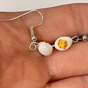 以下が含まれることがあります： ミニチュアのゆで卵をモチーフにした、ノベルティピアス。各ピアスは、銀色のイヤーフックに吊り下げられた、黄身が黄色い白い卵です。ピアスは半分に切られた卵のように見えるようにデザインされています。