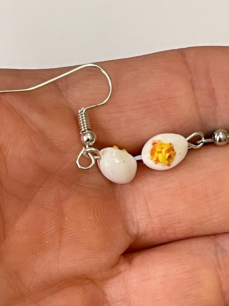 以下が含まれることがあります： ミニチュアのゆで卵をモチーフにした、ノベルティピアス。各ピアスは、銀色のイヤーフックに吊り下げられた、黄身が黄色い白い卵です。ピアスは半分に切られた卵のように見えるようにデザインされています。